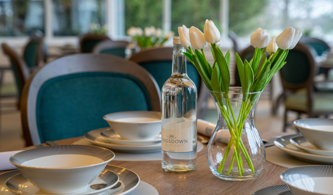 A dining table is laid with crockery with a glass bottle of water and vase of white tulips in the centre of the table, there's a glass bottle of water on table