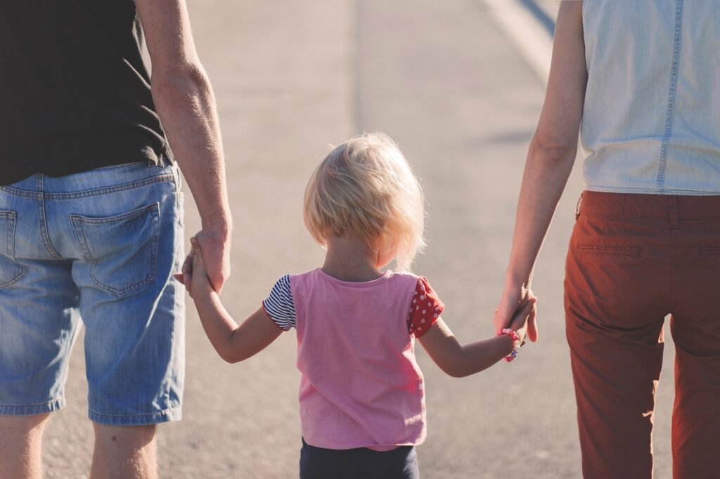 A toddler with a pink tshirt holds hands with two adults down a road. Only the backs of their heads and bodies can be seen.