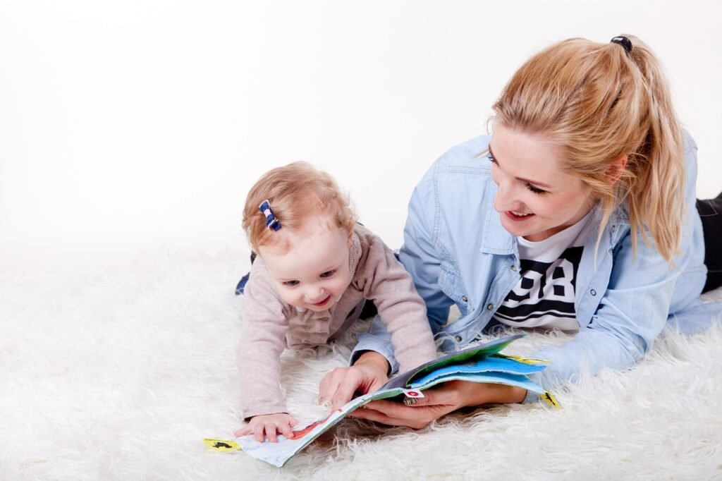A woman with a blue shirt lies on a fluffy blanket showing a toddler with a pink cardigan a children's book. They are both smiling.