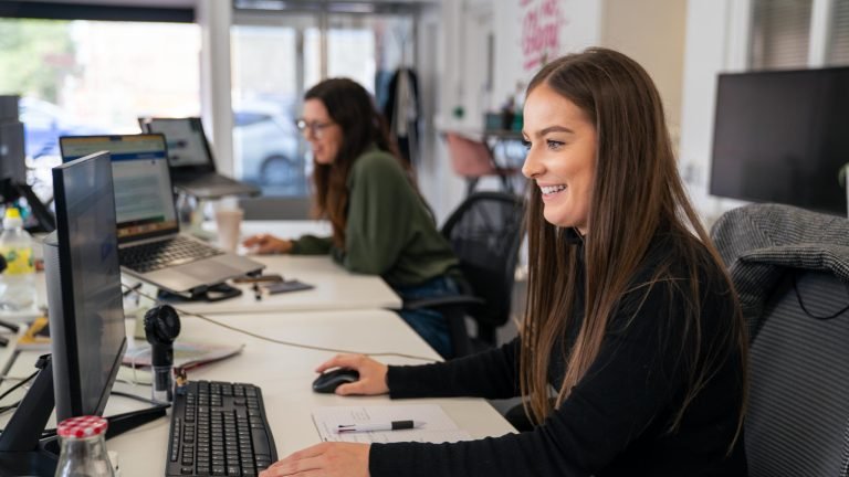 Two adults are working at at office desks at their computer screens