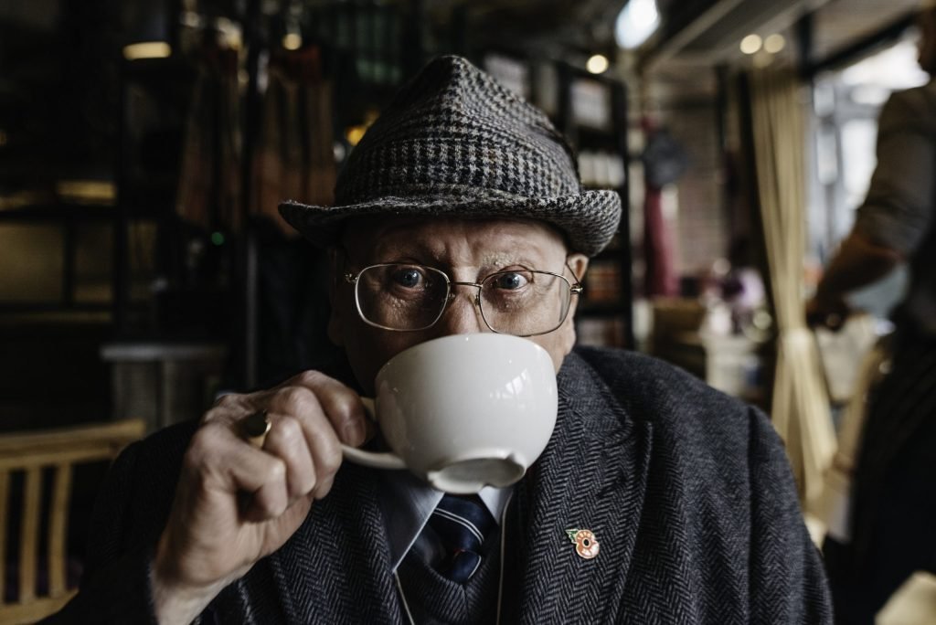 An old white man wearing a tweed jacket and hat sips from a coffee cup. He's wearing a signet ring and glasses, a tie and a poppy pin on his lapel
