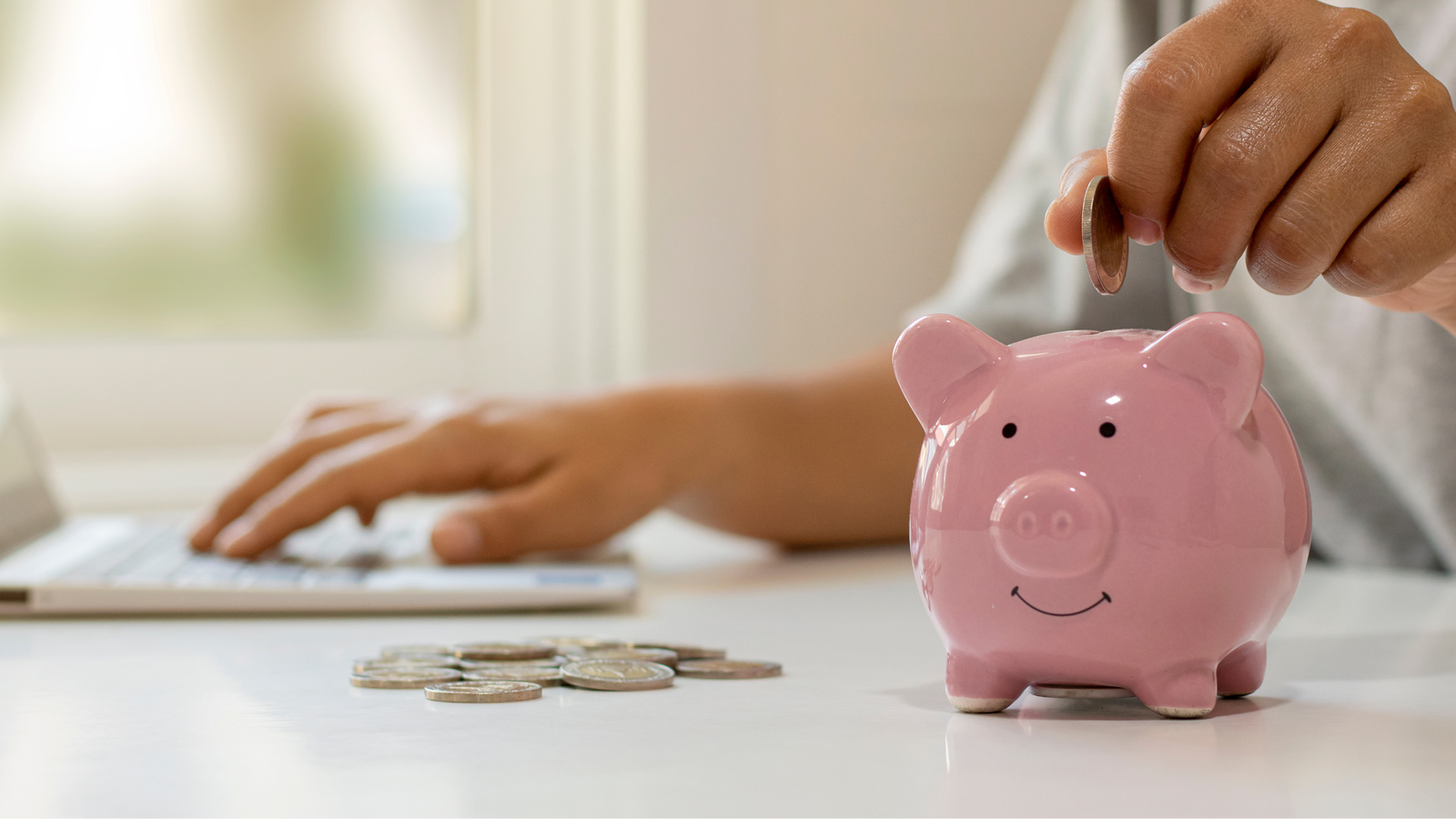 A person types on a laptop sharing marketing tips while placing a coin into a pink piggy bank on a table with scattered coins in the foreground.