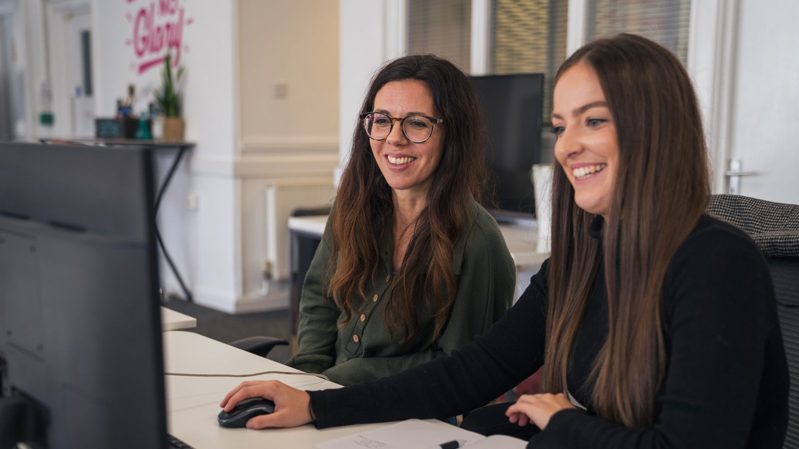 Two white women with long brown hair smiling at a computer screen.