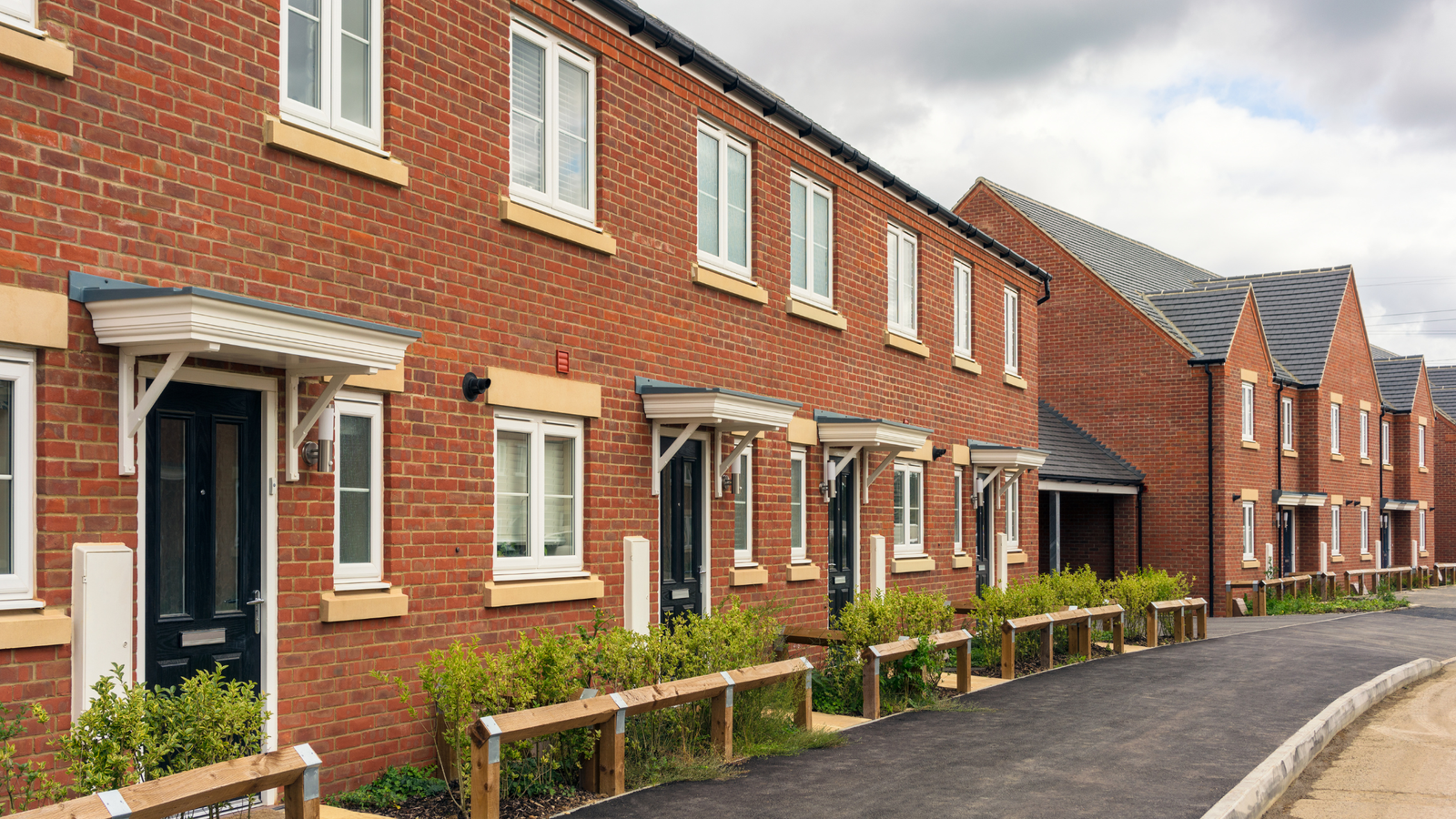 Row of newly built red brick terraced houses with black front doors, small porches and low hedges lining a paved pathway on a modern residential development.