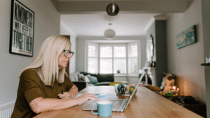 A white middle-aged woman with long blonde hair and glasses sits at a wooden dining table focused on working on a laptop.