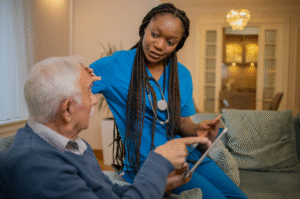 A Black care worker in blue scrubs supports an older white resident while looking at a tablet together in a warm, homely care setting.